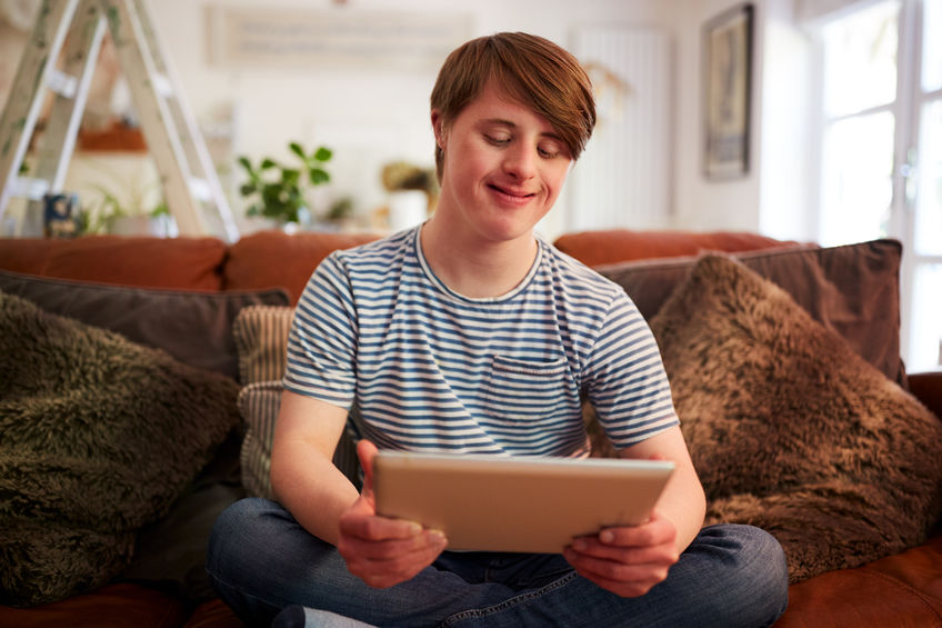 Young Downs Syndrome Man Sitting On Sofa Using Digital Tablet At Home