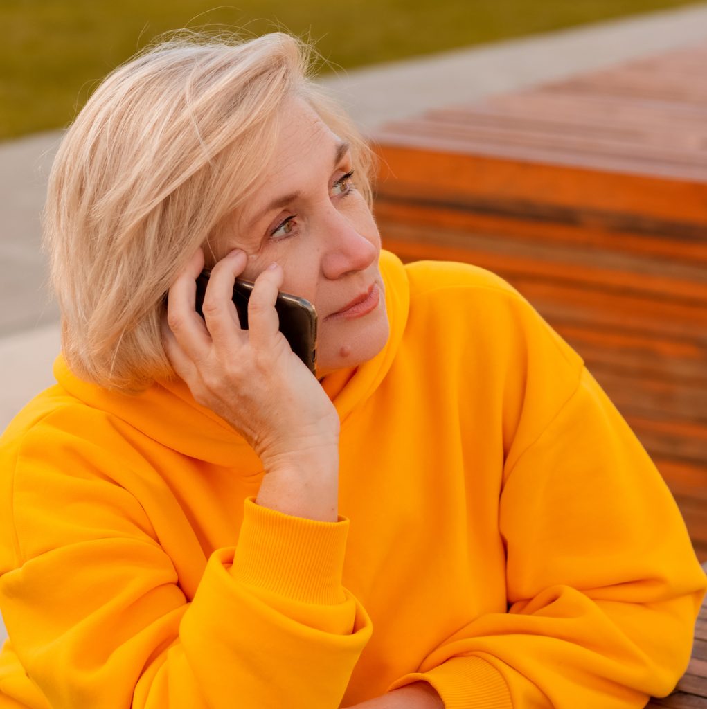 older woman talking on cellphone while sitting at a wooden table outside